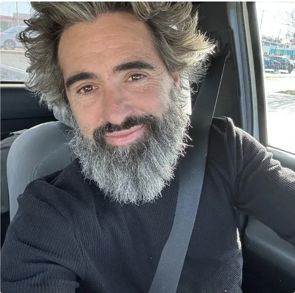 A bearded actor smiles in a car between takes