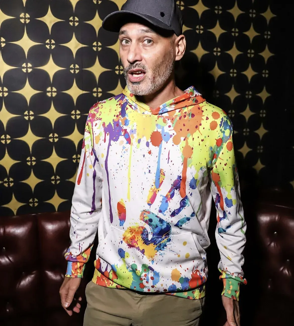 An actor in a paint-splatter hoodie in front of a gold patterned wall